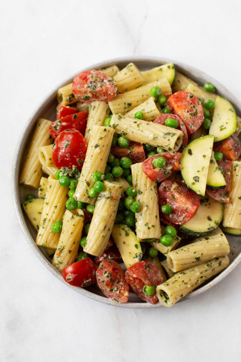 An overhead image of a bowl of creamy pesto pasta salad, made with fresh vegetables. It is sitting on a white marble surface.