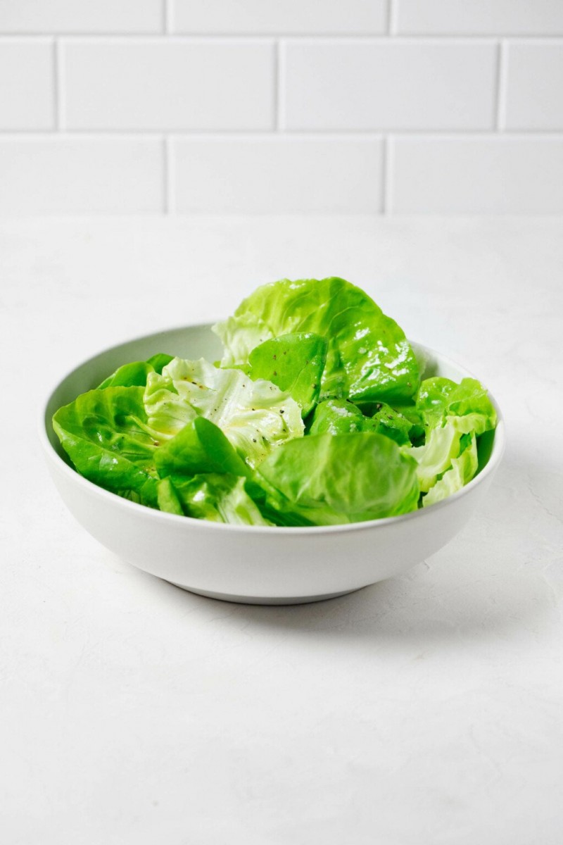 An angled image of a butter lettuce side salad, which is resting on a white surface. There are white tiles in the background.