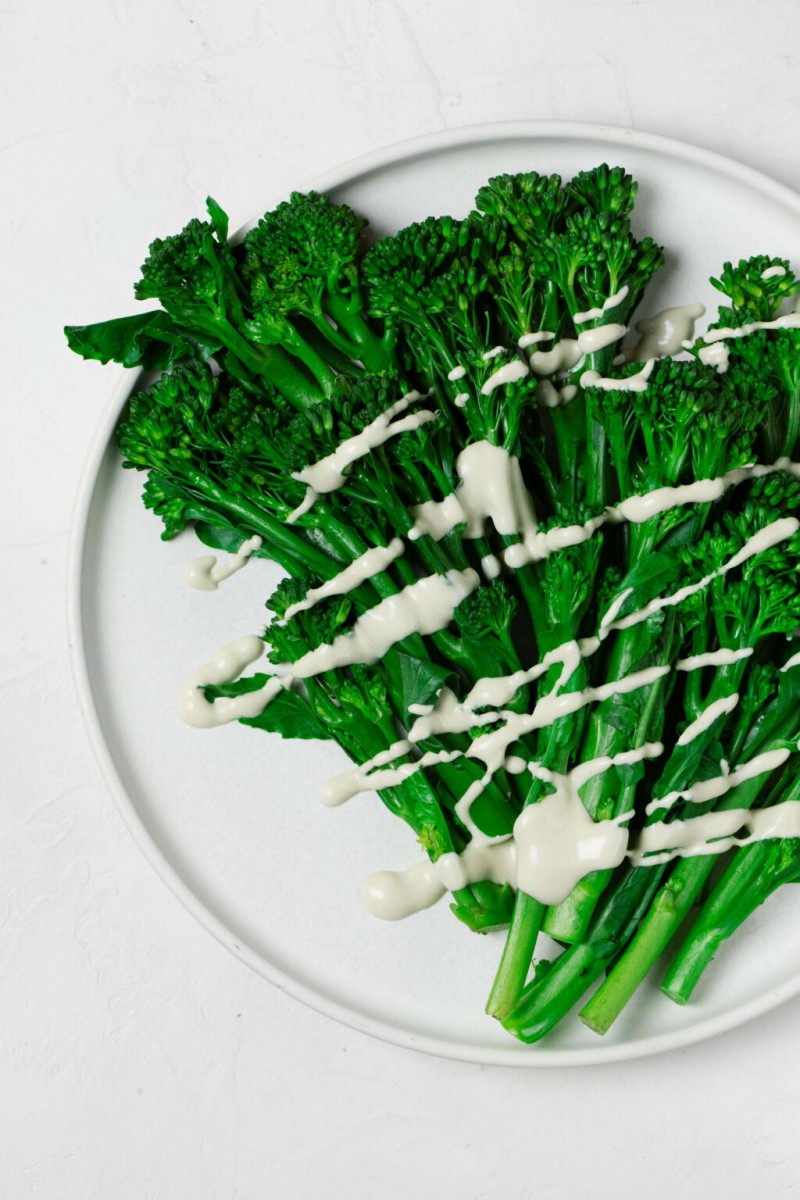 An overhead image of bright green, steamed broccolini resting on a white, round plate. The broccolini is drizzled with a creamy white tahini dressing.