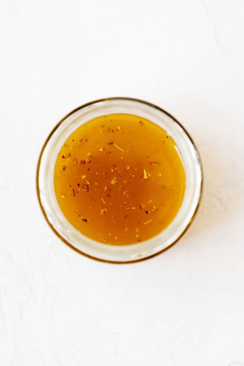 An overhead image of a clear, glass jar of homemade vinaigrette, resting on a white surface.