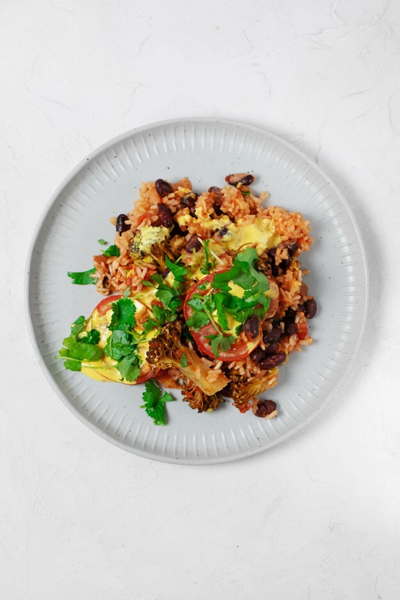 A photo of a round, grayish white plate, which has been topped by a bean and brown rice casserole. Green cilantro leaves are garnished on top.