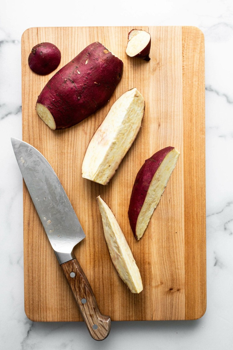 An overhead image of whole and sliced Japanese sweet potatoes on a light brown cutting board.