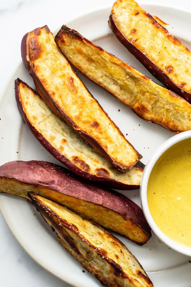 An overhead image of crispy roasted Japanese sweet potato wedges. They're arranged on a white platter with a bright yellow dipping sauce.