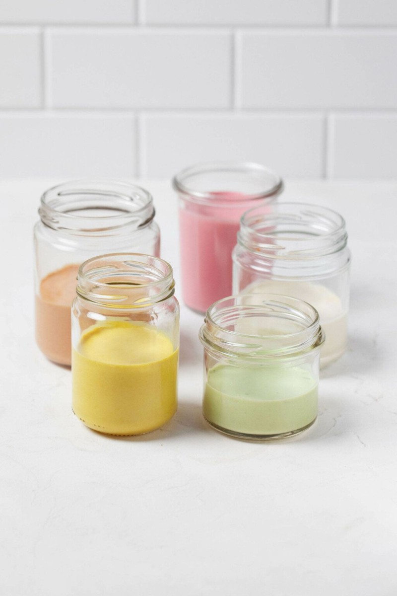 Five glass mason jars rest on a white surface. Each is filled with a different color of creamy tahini dressing.