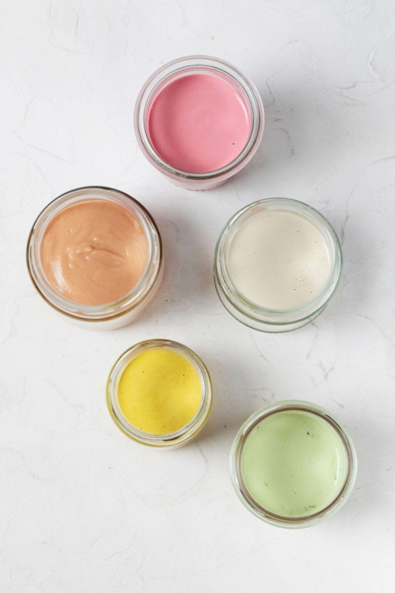 Five glass mason jars rest on a white surface. Each is filled with a different color of creamy tahini dressing.