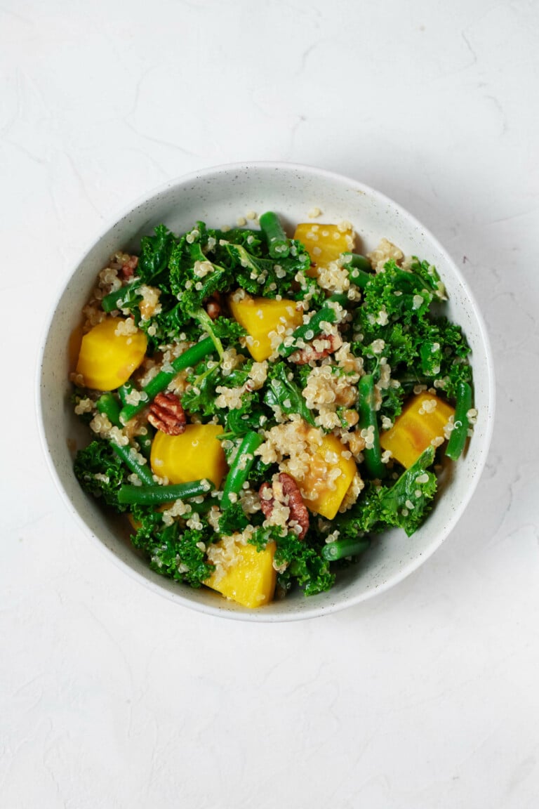 An overhead image of a white bowl, which is filled with a vibrantly-colored kale, golden beet, green bean, and quinoa salad. There are brown pieces of toasted pecan throughout.